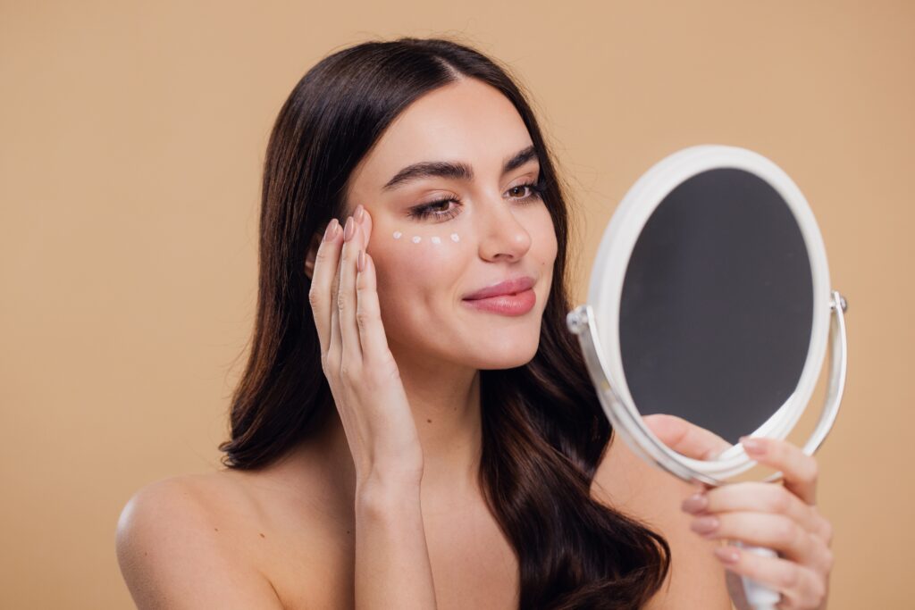 Woman gently applying eye cream under her eyes while looking in the mirror.
