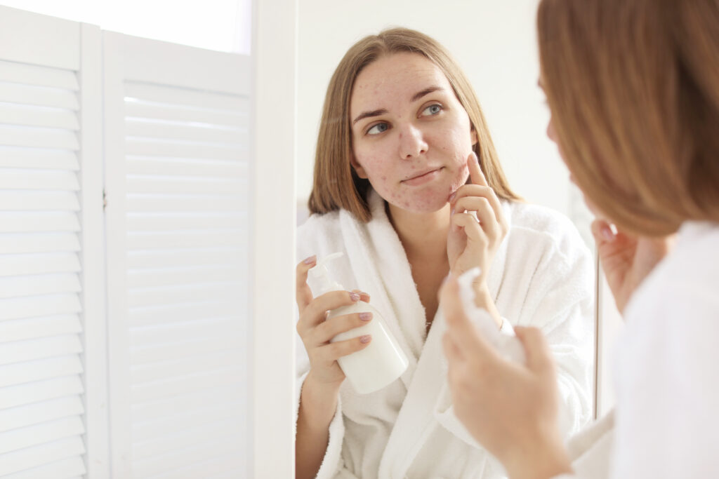 Woman looking in the mirror with dull, tired-looking skin while applying hydrating skincare.