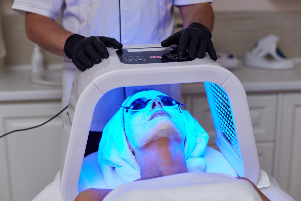 Client relaxing during an LED facial treatment while a professional light therapy device is positioned above her face.